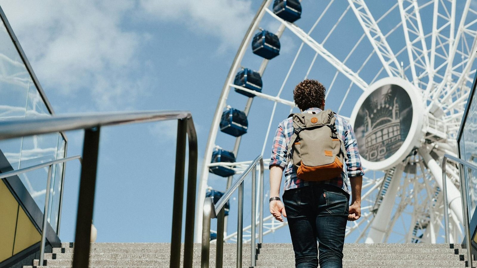 Back view of anonymous man in casual clothes with backpack walking up stairs leading to ferris wheel in daytime